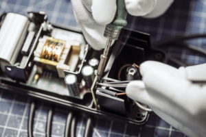 Nahaufnahme von behandschuhten Händen, die mit einem Lötkolben und einer Pinzette an elektronischen Komponenten im Inneren eines Geräts arbeiten. Der Arbeitsbereich ist mit einer gitterförmigen Matte versehen, was die Präzision im heiklen Montageprozess unterstreicht.