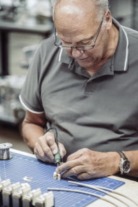 Ein Mann mit Brille und grauem Hemd ist konzentriert damit beschäftigt, an einer Werkbank Drähte zu löten. Er hält einen Lötkolben in der Hand und arbeitet an einer blauen Schneidematte, umgeben von verschiedenen Werkzeugen.
