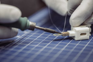 Nahaufnahme einer Person mit weißen Handschuhen, die ein kleines elektronisches Bauteil auf einer blauen, gittergemusterten Matte lötet. In einer Hand hält sie einen Lötkolben, mit der anderen Hand hält sie das Bauteil fest.