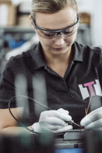 Eine Person mit Schutzbrille und Handschuhen arbeitet konzentriert mit Drähten und Werkzeugen. Sie trägt ein dunkles Poloshirt mit einem Logo und ist mit einer detaillierten Aufgabe beschäftigt, wahrscheinlich in einem elektronischen oder technischen Umfeld.