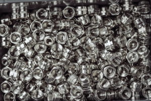 Ein Stapel glänzender, metallischer Muttern, meist silberfarben, füllt einen Behälter. Die Muttern haben innen ein Gewinde und einige haben außen geriffelte Stellen, was auf eine Verwendung in mechanischen oder Klempnerarbeiten hindeutet. Das Gesamtbild ist dicht gepackt und organisiert.
