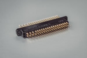 Nahaufnahme eines schwarzen elektrischen Steckers mit mehreren goldenen Pins. Der Stecker liegt auf einer glatten, hellgrauen Oberfläche, wodurch die lineare Anordnung der Pins und die komplizierten Details hervorgehoben werden.