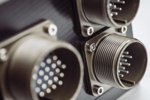 Nahaufnahme von drei Industriesteckverbindern, die auf einer schwarzen Platte montiert sind. Die Steckverbinder haben eine runde Form mit mehreren Metallstiften im Inneren. Sie bestehen aus einem metallischen Material und sind mit sichtbaren Schrauben befestigt.