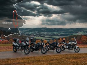 Fünf geparkte Motorräder auf einer asphaltierten Straße mit einer malerischen Kulisse aus sanften Hügeln und bewölktem Himmel. In der oberen linken Ecke ist eine Karte mit einer hervorgehobenen Route und „468 km“ eingeblendet, die einen umrissenen Weg zeigt.