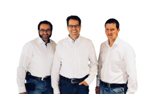 Drei Männer stehen da und lächeln in die Kamera. Sie tragen alle weiße Hemden und Jeans. Der Hintergrund ist schlicht weiß.