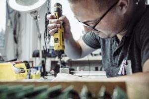 Eine Person mit Brille arbeitet mit einem gelben Akkuschrauber an einem kleinen Objekt an einer vollgestellten Werkbank. Im Hintergrund sind verschiedene Werkzeuge und Geräte zu sehen.
