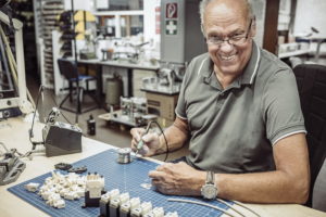 Ein lächelnder Mann in einem grauen Poloshirt sitzt an einer Werkbank voller elektronischer Bauteile und Werkzeuge. Er hält einen Lötkolben in der Hand und arbeitet an einer Platine. Der Raum sieht aus wie eine Werkstatt mit verschiedenen Geräten im Hintergrund.