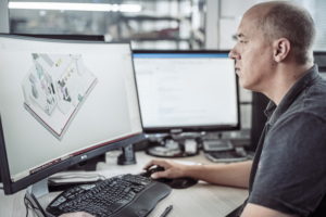 Eine Person sitzt an einem Schreibtisch und arbeitet an einem Computer, auf dessen Monitor ein CAD-Design angezeigt wird. Der Arbeitsbereich umfasst einen weiteren Monitor, eine Tastatur und verschiedene Büroartikel. Durch das Fenster fällt natürliches Licht in den Raum.