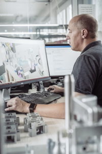 Eine Person sitzt an einem Schreibtisch und arbeitet an einem Computer mit zwei Monitoren. Auf dem Bildschirm wird eine detaillierte 3D-Modellierungssoftware angezeigt. Auf dem Schreibtisch sind verschiedene Metallteile angeordnet. Die Umgebung scheint ein Industrie- oder Ingenieurarbeitsplatz zu sein.