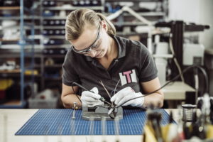 Eine Person mit Schutzbrille und Handschuhen lötet Drähte an einer Werkbank. Sie ist auf ihre Aufgabe konzentriert und in einer Werkstatt von Werkzeugen und Geräten umgeben. Auf dem Tisch liegt eine blaue Schneidematte.