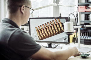 Eine Person mit Brille sitzt an einem Schreibtisch und blickt auf einen Computermonitor, auf dem ein vergrößertes Bild elektronischer Komponenten angezeigt wird. Neben dem Monitor steht ein digitales Mikroskop.