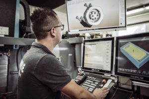 Eine Person bedient eine CNC-Maschine und konzentriert sich auf mehrere Monitore, auf denen 3D-Modelle und Schemata angezeigt werden. Die Person trägt eine Brille und ein graues Hemd in einer industriellen Umgebung.