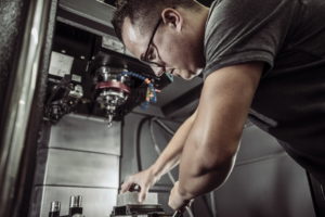 Eine Person mit Brille bedient eine CNC-Maschine und ist ganz auf ihre Arbeit konzentriert. Sie positioniert ein Metallstück unter dem Werkzeug der Maschine. Die Umgebung wirkt industriell, mit Metallkomponenten und Maschinen im Hintergrund.