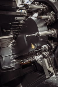 Nahaufnahme einer industriellen CNC-Maschine mit mehreren rotierenden Spindeln und Bohrern. Die Maschine ist aus Metall und hat verschiedene komplizierte Teile. Ein dreieckiges Warnsymbol ist sichtbar. Überall sind Späne verstreut, was auf aktive Nutzung hinweist.