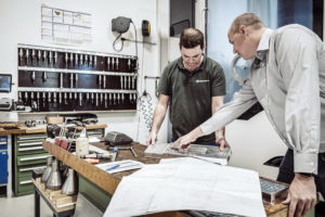 Zwei Männer in einer Werkstatt prüfen technische Dokumente, die auf einem Tisch ausgebreitet sind. Einer trägt ein grünes Hemd, der andere ein weißes. Im Hintergrund sind Werkzeuge auf einem Wandregal angeordnet. Im Vordergrund sind Konstruktionspläne zu sehen.
