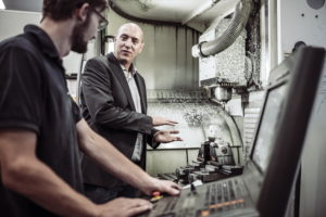 Zwei Männer in einer Fabrik, einer im Anzug, der andere im Poloshirt, unterhalten sich, während sie neben Industriemaschinen stehen. Der Mann im Anzug gestikuliert, während der andere ein Bedienfeld bedient. Im Hintergrund sind Metallspäne zu sehen.