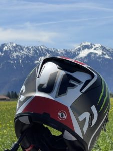 Ein Motorradhelm mit den Initialen „JF“ und verschiedenen Logos liegt auf einer grünen Wiese. Im Hintergrund sind schneebedeckte Berge und ein strahlend blauer Himmel mit vereinzelten Wolken zu sehen.