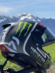 Eine Nahaufnahme eines schwarz-weißen Motorradhelms mit Monster Energy-Logo vor einer Kulisse aus schneebedeckten Bergen und blauem Himmel. Der Helm liegt auf einer Wiese mit verstreuten gelben Blumen.