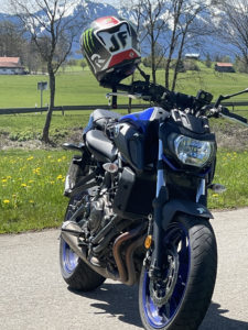 Ein blaues Motorrad mit einem Helm mit Visier und farbenfrohem Design steht auf einer Straße. Im Hintergrund sind grüne Felder, Bäume, Häuser und schneebedeckte Berge unter einem klaren blauen Himmel zu sehen.