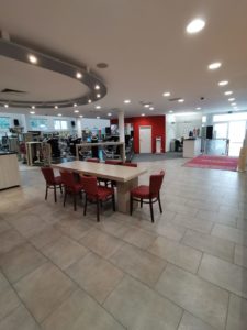 Ein geräumiger Fitnessraum mit Trainingsgeräten wie Laufbändern und Kraftmaschinen. Im Vordergrund steht ein Holztisch mit roten gepolsterten Stühlen. Der Boden ist gefliest und die Wände sind weiß und rot, große Fenster sorgen für natürliches Licht.
