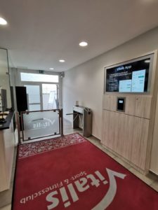 Eingang zum Fitnesscenter mit einer roten Matte mit der Aufschrift „Vitalis“. In der Mitte ein Glastor und ein Drehkreuz, rechts ein digitaler Bildschirm mit App-Informationen. Helles, modernes Interieur mit hellen Holzakzenten und Deckenbeleuchtung.