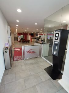 Eine moderne Banklobby mit Drehkreuzeingang, Selbstbedienungsautomat und Rezeptionsbereich. Der Innenraum ist in Rot und Weiß gehalten und verfügt über geflieste Böden und helle Beleuchtung. Glaswände trennen die verschiedenen Bereiche des Raums.