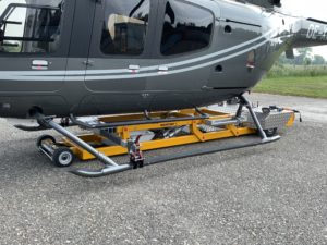 Ein am Boden geparkter Hubschrauber mit darunter angebrachter gelber Wartungsausrüstung mit Rädern und einem Metallrahmen. Das Flugzeug ist grau mit stromlinienförmigem Design und Fenstern und steht auf einer Kiesfläche mit Gras im Hintergrund.