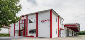 Ein modernes weißes Gebäude mit roten Akzenten und einem „SHERPA“-Logo auf der Fassade, mit mehreren Fenstern und einer gepflasterten Auffahrt. Links ist ein Baum und darüber ein bewölkter Himmel.