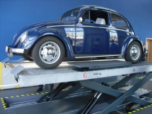 Ein blauer Oldtimer steht drinnen auf einer hydraulischen Hebebühne. Die Hebebühne weist Gewichtsgrenzen von 3500 kg und 4200 kg auf. Der Bereich ist mit gelb-schwarzem Absperrband markiert.