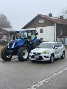 Ein blauer Traktor steht neben einem weißen SUV in einer Vorstadtstraße. Auf der Seite des SUV steht ein Text und beide Fahrzeuge stehen an einem nebligen Tag mit einem großen Haus und Bäumen im Hintergrund.