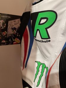 Nahaufnahme der Motorradrennausrüstung mit dem grünen „R Fahrschule“-Logo und einem Monster Energy-Emblem. An der Wand hängt außerdem ein kleines Poster mit Motorradgrafiken und Autogrammen, darunter eine Notiz mit der Aufschrift „Für Ruben“.