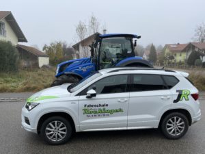 Ein weißer Fahrschul-SUV mit grünen Aufklebern steht neben einem großen blauen Traktor auf einer Straße. Im Hintergrund sind an einem nebligen Tag Wohnhäuser und Bäume zu sehen.
