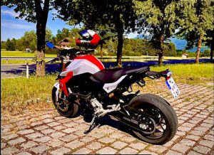 Ein rot-weißes Motorrad mit einem Helm auf dem Sitz steht auf einem gepflasterten Platz. Im Hintergrund sind Bäume, Gras und ein klarer blauer Himmel zu sehen, was eine ländliche oder malerische Umgebung suggeriert.