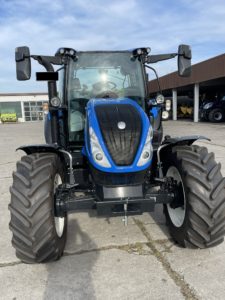 Ein blau-schwarzer Traktor mit großen Reifen steht auf einer Betonfläche vor einem Industriegebäude. Der Himmel ist teilweise bewölkt. Der Traktor hat sichtbare Seitenspiegel und ein schlankes, modernes Design.