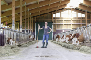 Eine Person steht selbstbewusst in einem offenen Bauernhaus und hält einen Besen in der Hand. Auf beiden Seiten sind Rinder zu sehen, die in Ställen grasen. Sonnenlicht erhellt die Holzbalken und das moderne Innere der Scheune.