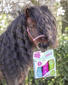 Ein Pony mit langer, lockiger Mähne steht draußen zwischen blühenden Pflanzen und hält ein Set mit Magic Brush-Pflegewerkzeugen im Maul. Das Set ist bunt und enthält rosa, blaue und grüne Bürsten. Das Pony hat ein rosa Halfter aus Seil.