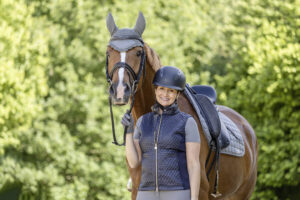 Eine Person mit Helm und Reitausrüstung steht neben einem braunen Pferd mit Sattel und Zaumzeug. Das Pferd trägt eine Schutzhülle an den Ohren. Sie stehen draußen vor einem Hintergrund aus üppigen grünen Bäumen und sehen beide zufrieden aus.