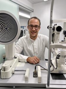 Ein Mann mit Brille und weißem Hemd sitzt in einer modernen Klinik an einem Augenuntersuchungsgerät. Er lächelt in die Kamera. Der Raum ist hell beleuchtet und wirkt elegant und professionell.