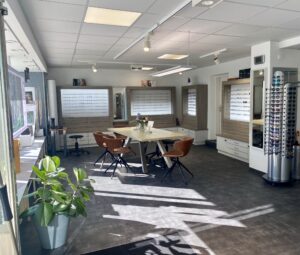 Helles Interieur eines Optikerladens mit Brillen auf Wandregalen und einem zentralen Tisch mit Stühlen. Große Fenster lassen natürliches Licht in den Raum. Eine Topfpflanze und Dekorationsstücke werten das Ambiente auf.