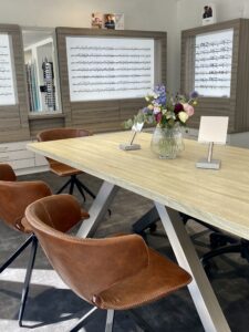 In der Praxis eines Optikers steht ein Holztisch, der von braunen Lederstühlen umgeben ist. Im Hintergrund sind Vitrinen mit verschiedenen Brillen ausgestellt. Auf dem Tisch steht eine durchsichtige Vase mit bunten Blumen, zusammen mit kleinen Ausstellungsständern.