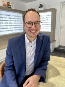 Ein Mann mit Brille und blauem Blazer lächelt in der Praxis eines Optikers in die Kamera. An der Wand hinter ihm hängen Reihen von Brillengestellen.