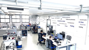 Ein helles, modernes Labor mit mehreren Mitarbeitern an Schreibtischen und Computern. An den Wänden sind große Schriftzüge mit Bezug zur Laborarbeit zu sehen. Geräte und Werkzeuge sind in dem sauberen, übersichtlichen Raum verteilt.