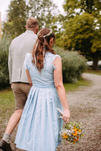 Eine Frau in einem hellblauen Kleid, die einen Blumenstrauß in der Hand hält, geht neben einem Mann in traditioneller Tracht einen von Grün umgebenen Schotterweg entlang.