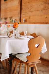 Ein Esstisch aus Holz mit einer weißen Tischdecke, gedeckt mit Gläsern, Tellern, Besteck und Blumengestecken als Tischdekoration. Um den Tisch herum stehen Holzstühle mit herzförmigen Ausschnitten.